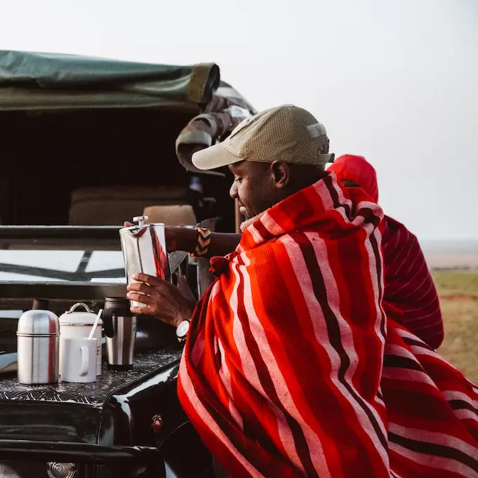 Guest wrapped in red blanket enjoying beverage at safari vehicle in open landscape, Speke’s Camp, Maasai Mara, Kenya