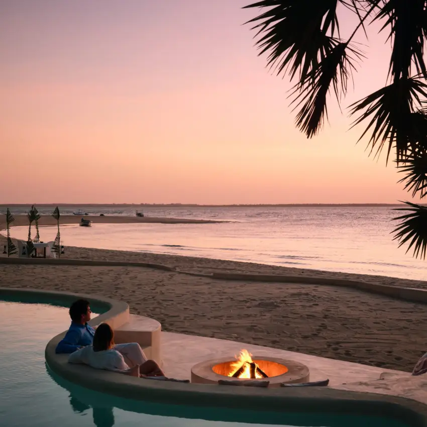 Couple relaxing in infinity pool by firepit overlooking beach at sunset, The Cabanas, Lamu Archipelago, Kenya