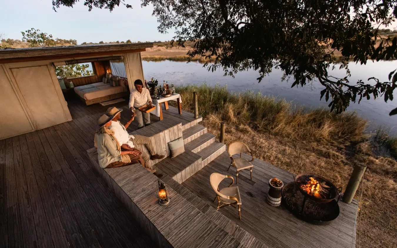 Guests relaxing by firepit on wooden deck overlooking waterhole at dusk, Savuti Camp, Linyanti Wildlife Reserve, Botswana