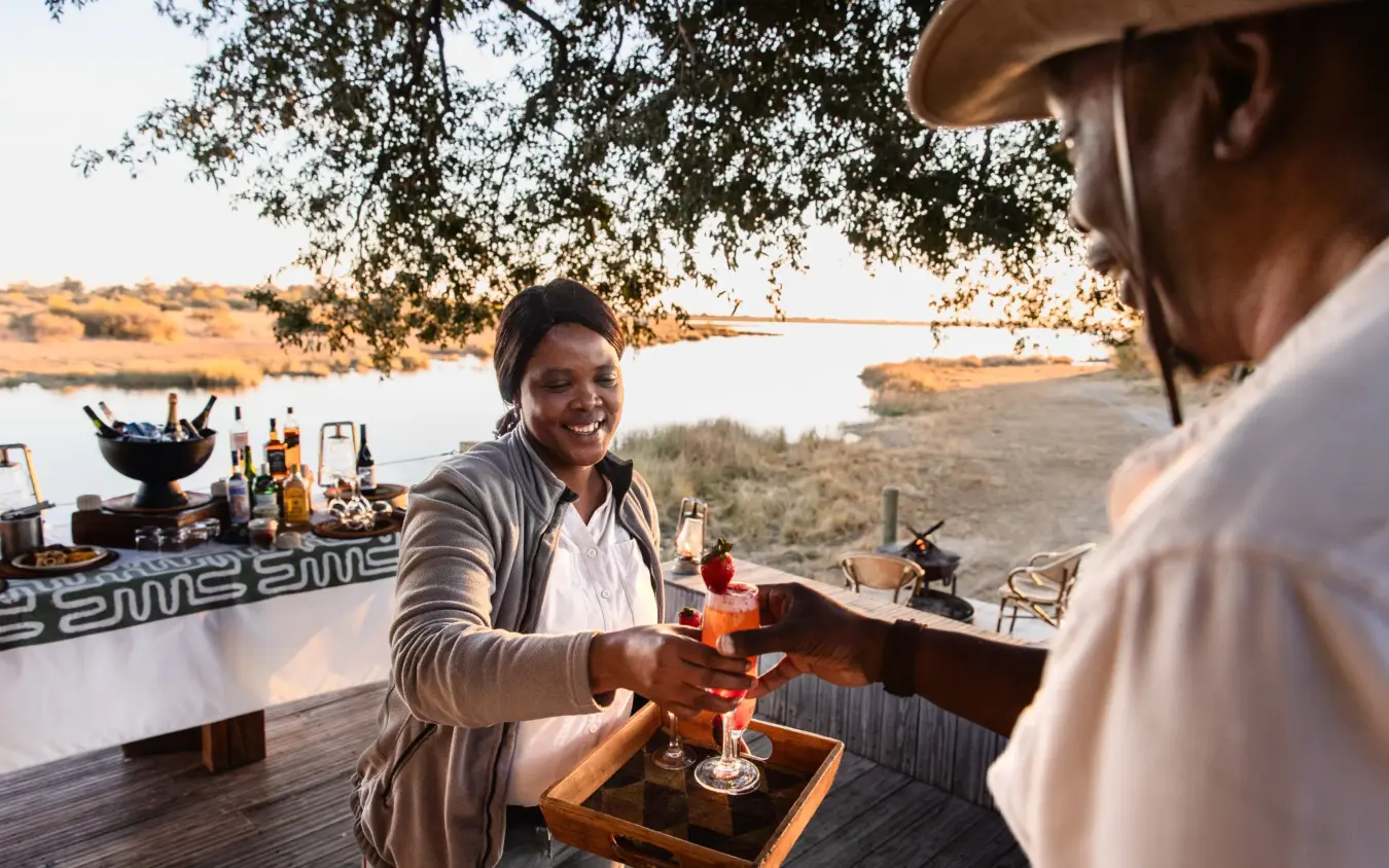 Server pouring drinks at sunset deck overlooking riverside landscape, Savuti Camp, Linyanti Wildlife Reserve, Botswana