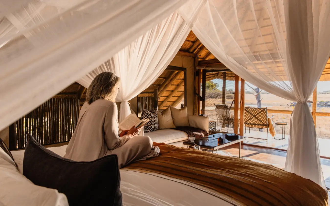 Guest reading on bed in safari tent with sheer curtains overlooking savanna, Savuti Camp, Linyanti Wildlife Reserve, Botswana